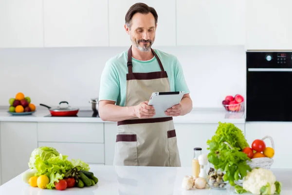 Meraklı, zeki, sakallı bir adamın tablet tutarken çekilmiş fotoğrafı. Yemek tarifi, mutfak önlüğü, kapalı bir evde mutfak önlüğü. — Stok fotoğraf