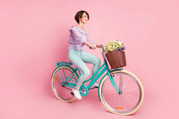 Foto lateral de perfil de tamanho completo da jovem linda menina sonhadora andando de bicicleta com flores na cesta isolada no fundo de cor rosa — Fotografia de Stock