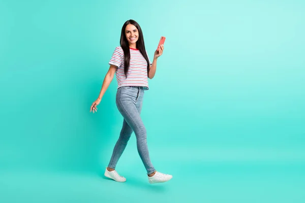 Full storlek foto av trevlig brunett optimistisk lång frisyr dam gå hålla telefon slitage t-shirt jeans sneakers isolerade på teal färg bakgrund — Stockfoto