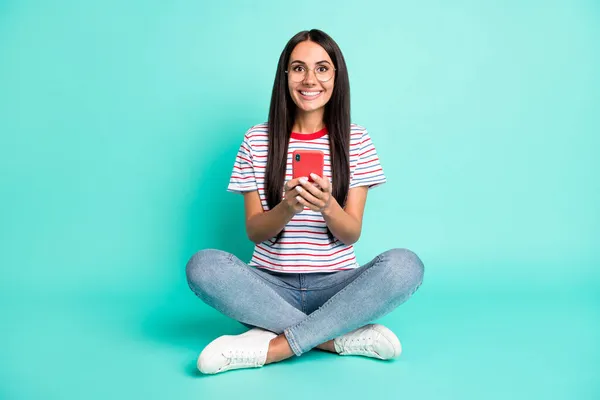 Full storlek foto av optimistisk brunett frisyr dam sitta skriva telefon slitage t-shirt jeans sneakers isolerad på teal bakgrund — Stockfoto