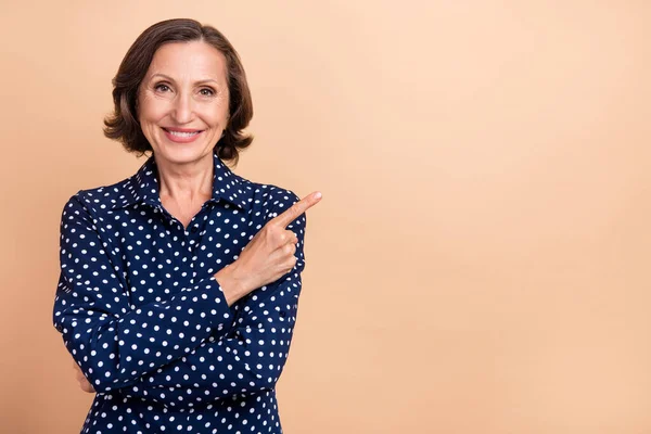 Foto van aantrekkelijke mooie oudere vrouw dragen gestippelde shirt wijzende vinger lege ruimte glimlachen geïsoleerde beige kleur achtergrond — Stockfoto