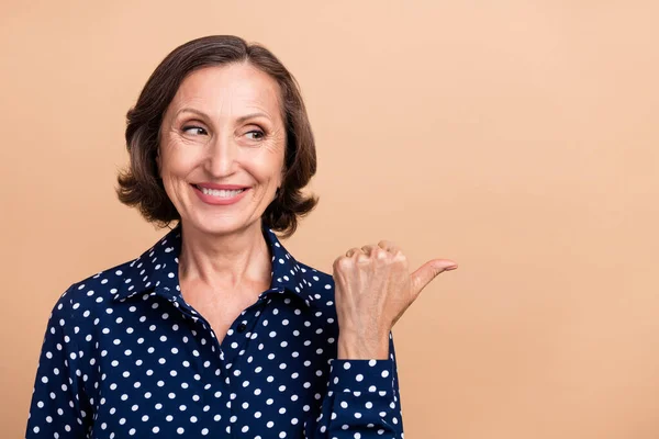 Fénykép csinos fényes idős nő viselni pontozott ing keres mutató ujj üres tér mosolygó elszigetelt bézs színű háttér — Stock Fotó