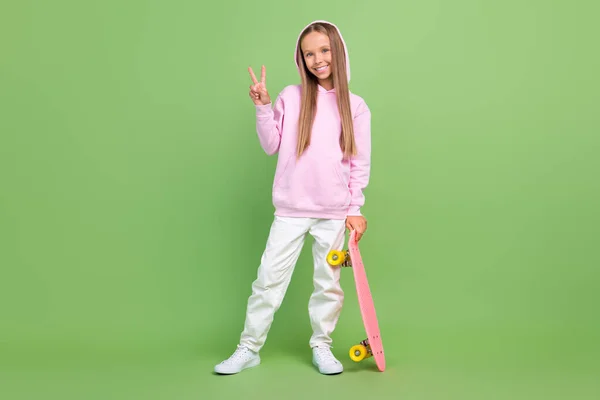 Full size photo of funny small blond girl hold skate show v-sign wear hoodie trousers sneakers isolated on green background — Stock Photo, Image