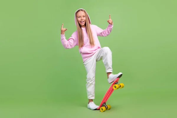 Foto de longitud completa de la pequeña chica rubia fresca paseo skate show rock wear sudadera con capucha pantalones zapatillas aisladas sobre fondo verde — Foto de Stock