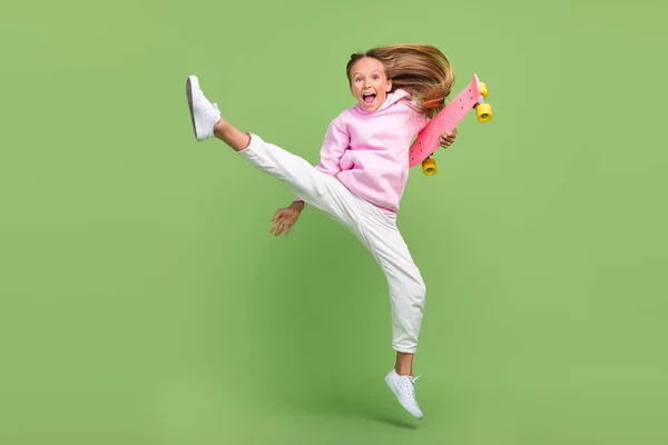 Ganzkörper-Foto von coolen kleinen blonden Mädchen springen mit Skate tragen Kapuzenpullover Hose Turnschuhe isoliert auf grünem Hintergrund — Stockfoto