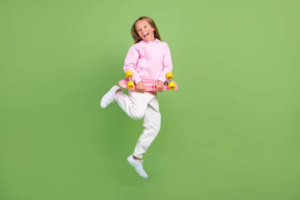 Foto de longitud completa de divertido salto niña rubia con skate desgaste sudadera con capucha pantalones zapatillas aisladas sobre fondo verde —  Fotos de Stock