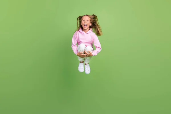 Foto de tamaño completo de cool niña rubia salto usar sudadera pantalones zapatillas aisladas sobre fondo verde — Foto de Stock