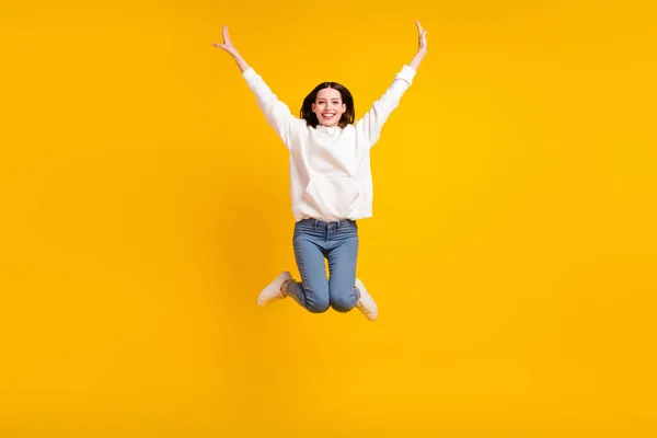 Photo of funky cheerful lady jump raise hands wear white hoodie jeans footwear isolated yellow color background — Stock Photo, Image