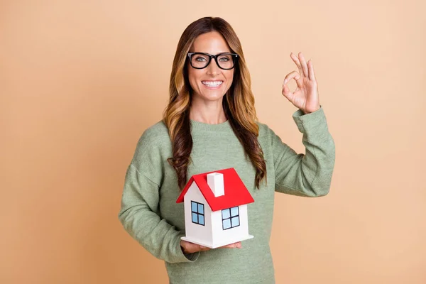 Portré vonzó vidám nő holding house figura mutatja ok-jel elszigetelt bézs pasztell szín háttér — Stock Fotó