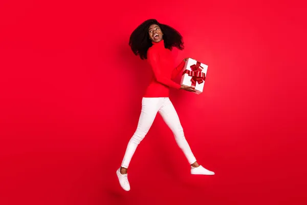 Tamaño completo perfil lado foto de la joven afro mujer feliz sonrisa positiva mantenga presente caja regocijo salto aislado sobre fondo de color rojo — Foto de Stock