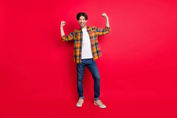 Photo de jeune homme assez fort habillé vêtements à carreaux montrant les muscles du bras souriant isolé fond de couleur rouge — Photo