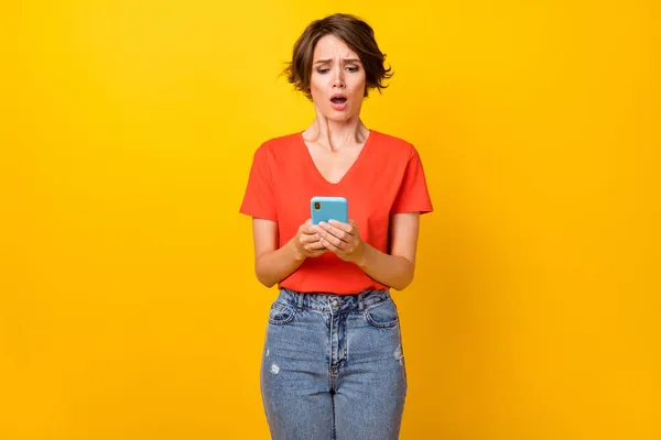 휴대폰을 들고 있는 슬픈 갈색 머리 여인의 사진은 선명 한 노란색 배경에 고립된 주황색 티셔츠를 입고 있다 — 스톡 사진