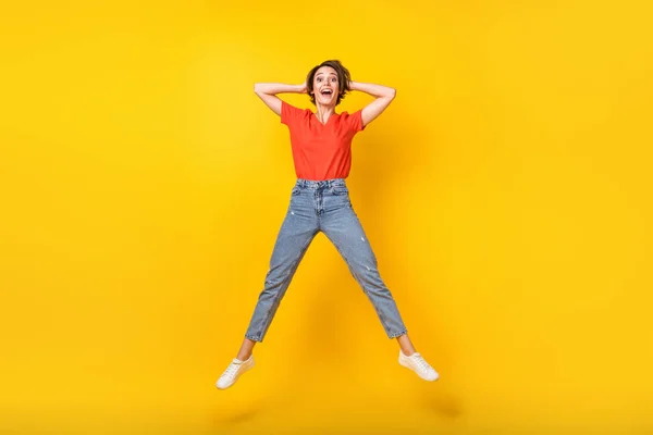 Foto de tamaño completo de la fresca morena optimista dama salto manos cabeza desgaste rojo camiseta jeans zapatillas aisladas sobre fondo de color amarillo —  Fotos de Stock