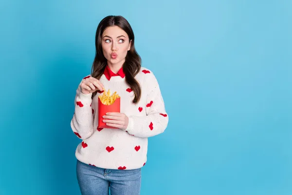 Foto av ung kvinna klädd hjärtan tryck pullover äta pommes frites läppar skrek ser tomt utrymme isolerad blå färg bakgrund — Stockfoto