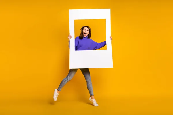Foto de encantadora dulce mujer joven usar suéter púrpura saltar sosteniendo marco de foto blanco aislado color naranja fondo — Foto de Stock