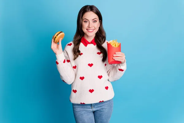 Fénykép bájos csinos fiatal hölgy viselni szív nyomtatott pulóver eszik gyorsétterem burgonya burger elszigetelt kék szín háttér — Stock Fotó