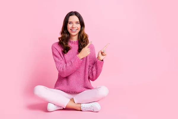 Full size photo of optimistic nice brunette lady sit point empty space wear pink sweater trousers sneakers isolated on pink background — Stock Photo, Image