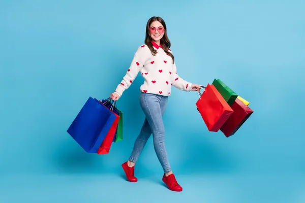 Teljes hosszúságú fotó fiatal nő öltözött szív nyomtatás pulóver szemüveg séta karok bolt táskák elszigetelt kék szín háttér — Stock Fotó