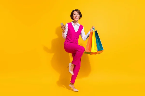 Photo of adorable lucky young woman dressed pink suit holding shopping bags modern gadget isolated yellow color background — Stock Photo, Image