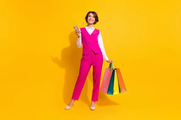 照片上，甜美甜美的年轻女子穿着粉色西服，抱着购物者的现代小玩意，看着空旷的空间，孤立的黄色背景 — 图库照片