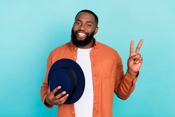 Fotografie mladého černocha šťastný pozitivní úsměv show mír cool V-sign hold klobouk izolované přes tyrkysové barvy pozadí — Stock fotografie