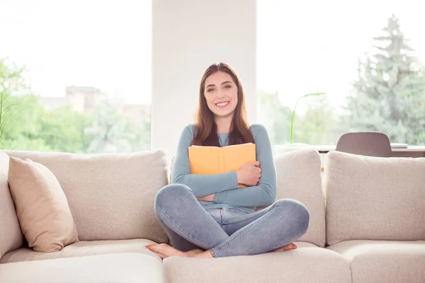 Полная длина фото молодой привлекательной женщины счастливой позитивной улыбкой обнимашки книги сидеть лотос поза диван — стоковое фото
