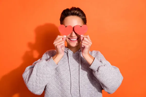 Φωτογραφία από αστείο γλυκό νεαρή γυναίκα ντυμένος γκρι κουκούλα μικρές κόκκινες καρδιές καλύπτουν τα μάτια χαμογελώντας απομονωμένο φόντο πορτοκαλί χρώμα — Φωτογραφία Αρχείου