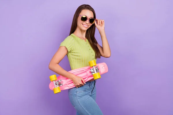 Retrato de menina muito alegre tocando especificações segurando skate posando descanso fim de semana isolado sobre roxo cor violeta fundo — Fotografia de Stock