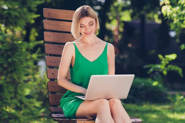 Фото блестящей очаровательной молодой леди носить зеленый наряд улыбаясь сидя скамейка печатая современный гаджет за пределами городской улицы — стоковое фото