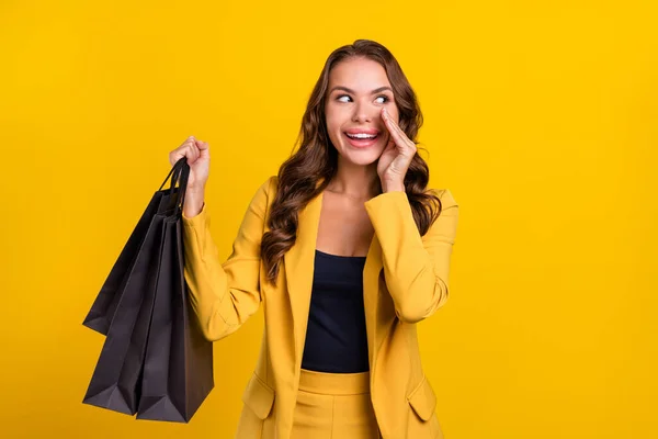 Porträtt av ganska glada vågiga hår kvinna håller väskor säger nyheter promo isolerad över ljusa gula färg bakgrund — Stockfoto