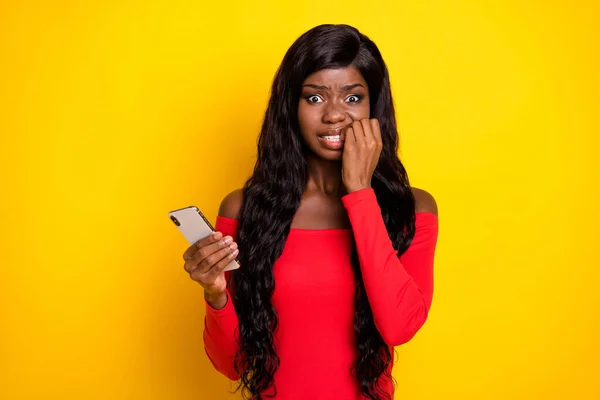 Photo of sweet scared dark skin lady wear off-shoulders top holding modern device biting fingers isolated yellow color background.