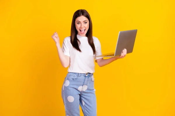 Fénykép a fiatal lány boldog pozitív mosoly hold laptop öröm győzelem ököl kéz elszigetelt sárga szín háttér — Stock Fotó