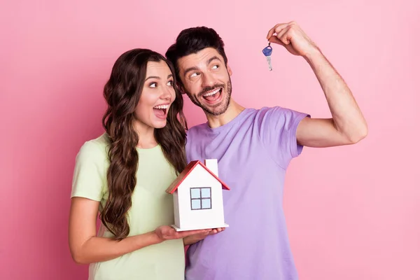 Retrato de atractiva pareja alegre asombrada abrazando la celebración de crédito pequeña llave en casa aislado sobre fondo de color pastel rosa —  Fotos de Stock