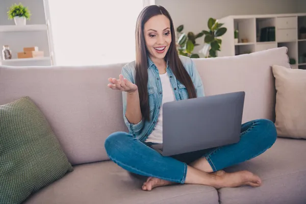 Retrato de atractiva mujer de pelo largo alegre y alegre sorprendido sentado en diván utilizando el ordenador portátil que llama a vídeo en casa en interiores —  Fotos de Stock