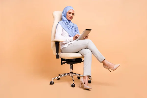 Full size photo of young arabic modern lady type tablet sit wear headscarf shirt eyewear pants stilettos isolated on beige background — Stock Photo, Image