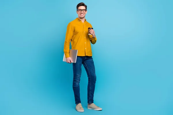 Foto av positiv avslappnad kille kaffe paus håll bärbar dator bära gul skjorta jeans skor isolerad blå färg bakgrund — Stockfoto