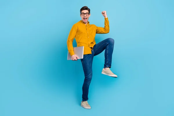 Foto van kampioen jongen houden pc vieren overwinning dragen gele shirt jeans sneakers geïsoleerde blauwe kleur achtergrond — Stockfoto
