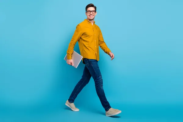 Full storlek profil foto av roliga brunett boss man hold laptop gå bära gul skjorta glasögon jeans skor isolerad på blå färg bakgrund — Stockfoto