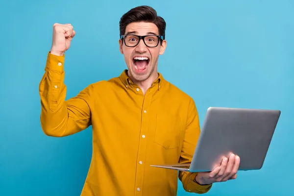 Foto van verbaasd gek kerel houden laptop schreeuwen verhogen vuist slijtage specs geel shirt geïsoleerde blauwe kleur achtergrond — Stockfoto