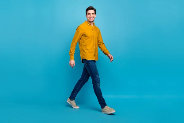 Foto de chico guapo positivo alegre caminar ir a usar pantalones vaqueros camisa amarilla zapatillas aisladas de color azul de fondo — Foto de Stock