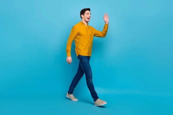 Profilfoto von positiven netten Kerl zu Fuß Straße Welle Hand tragen gelb Hemd Jeans Turnschuhe isoliert blaue Farbe Hintergrund — Stockfoto