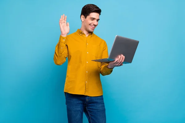Foto di socievole amichevole ragazzo tenere laptop onda mano remoto video chiamata usura giallo camicia isolato blu colore sfondo — Foto Stock