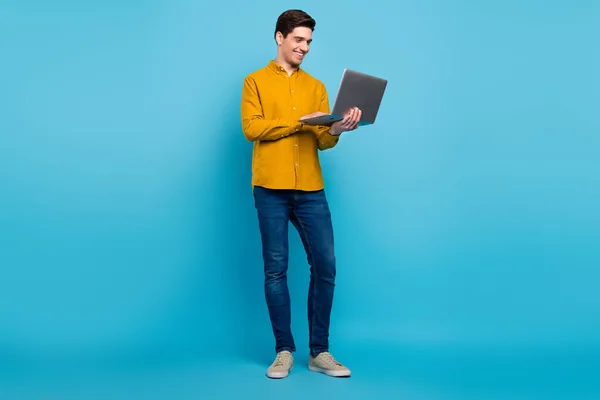 Full size foto van slimme brunet man look laptop dragen geel shirt jeans schoenen geïsoleerd op blauwe kleur achtergrond — Stockfoto