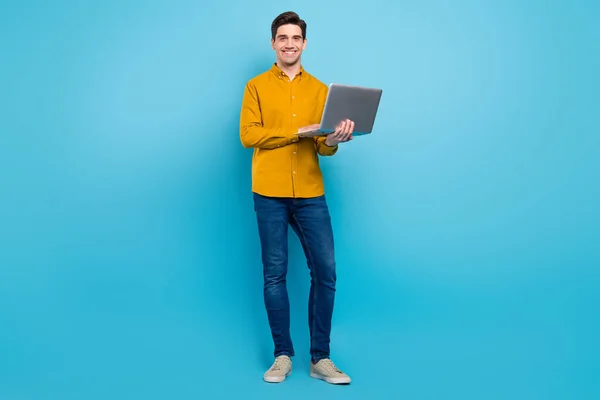 Foto van vrolijke freelancer kerel houden netbook typen dragen geel shirt jeans schoenen geïsoleerd blauw kleur achtergrond — Stockfoto