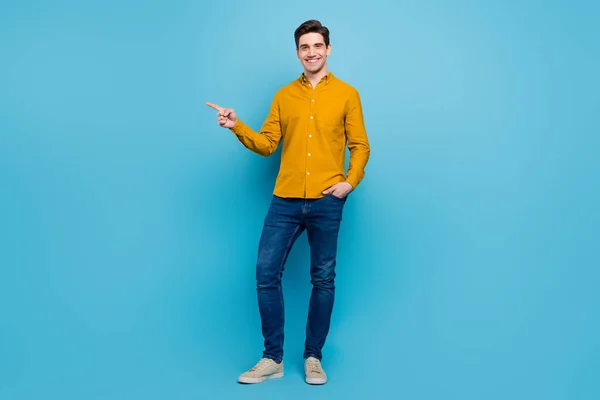 Foto av promotor killen ange pekfinger tomt utrymme bära gul skjorta jeans skor isolerad blå färg bakgrund — Stockfoto
