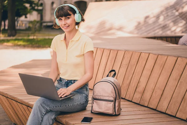 Photo de jeune femme heureuse et agréable écouter de la musique tenir ordinateur portable assis banc sourire à l'extérieur ville en plein air — Photo