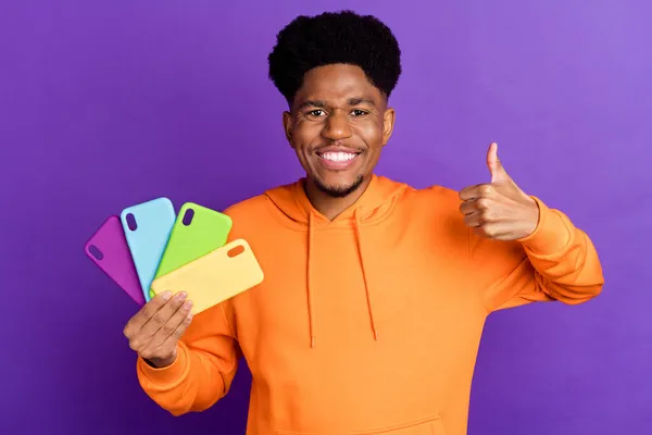 Foto di bruna afro americano uomo make thumb up hold phone case indossare felpa con cappuccio maglione arancione isolato su sfondo di colore viola — Foto Stock