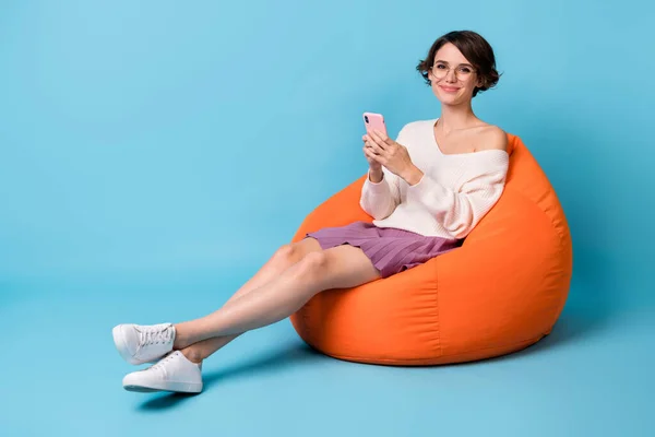 Foto retrato vista de corpo inteiro da mulher segurando telefone em duas mãos sentadas em cadeira de beanbag laranja isolado no fundo de cor azul pastel — Fotografia de Stock