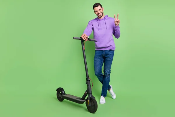 Foto de cuerpo completo del joven alegre hombre feliz sonrisa positiva paseo scooter mostrar la paz v-signo aislado sobre fondo de color verde —  Fotos de Stock