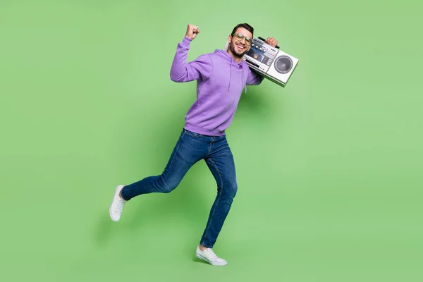 Full size photo of funky millennial brunet guy hold boom box dance wear eyewear hoodie sneakers isolated on green background — стоковое фото